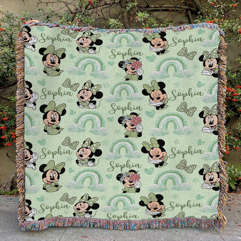 May include: A light green throw blanket featuring a repeating pattern of Minnie Mouse, rainbows, and butterflies. The name "Sophia" is written in cursive throughout the design. The blanket has a fringed edge.