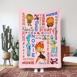 May include: A pink blanket with cartoon characters and the word "Noxie" in different colours and fonts. The blanket has a grid pattern and features several illustrated figures with unique designs. The blanket is held up by a person wearing white shoes.