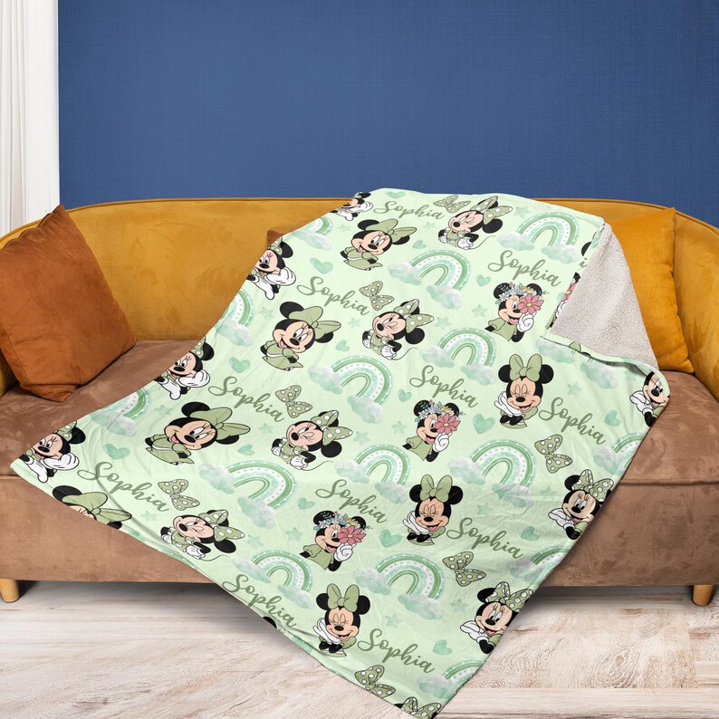 May include: A light green blanket featuring a pattern of Minnie Mouse characters, rainbows, and butterflies. The name "Sophia" is written throughout the design. The blanket is draped over a brown couch.