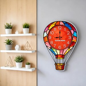 May include: A hot air balloon-shaped wall clock with a red face and black hands. The clock's face has white numbers and the text "Custom Name". The balloon design features colorful stained glass-style panels. The clock is mounted on a gray wall.