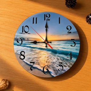 Op de afbeelding: Ronde wandklok met een strandscène met een blauwe lucht, oceaangolven en een zonsondergang. De klok heeft zwarte cijfers en wijzers, met een rode secondewijzer. De klok staat op een houten oppervlak.
