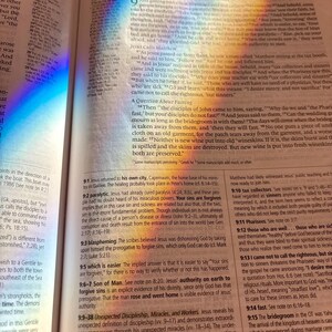 Może przedstawiać: Otwarta Biblia z tekstem, oświetlona tęczą światła. Strony ukazują drukowane słowa. Światło tworzy kolorowy efekt na stronach, podkreślając tekst. Książka leży na drewnianej powierzchni.