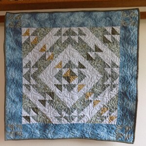 May include: A square fabric quilt featuring a central diamond pattern composed of white, sage green, and mustard yellow triangles. The quilt is framed by a blue border with a swirling stitched texture and dark gray binding along the edges.