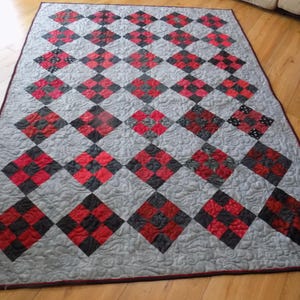 May include: A rectangular quilt featuring a repeating pattern of diamond-shaped blocks. Each diamond consists of a four-patch grid of red and black fabric squares. The background fabric is light gray with a swirling, quilted texture.