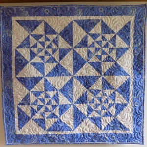 May include: A handmade quilt featuring a geometric pattern of blue and white triangles. The quilt has a blue border with a floral design and intricate quilting throughout. The overall design is symmetrical and visually appealing.