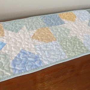 May include: A rectangular quilted table runner featuring a repeating star pattern in soft pastel shades of light blue, pale yellow, and sage green. The fabric has a textured, wavy stipple quilting design throughout and a light green border.