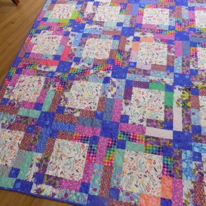 May include: A rectangular patchwork quilt featuring a repeating grid of white fabric squares printed with colorful floral and botanical patterns, interspersed with vibrant strips of blue, pink, purple, green, and rainbow-colored fabric blocks.