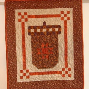 May include: A brown and orange quilted wall hanging with a design of an acorn. The acorn is surrounded by a double orange frame with a white background.