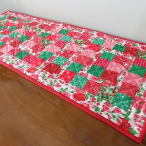 Può includere: Un runner da tavola trapuntato rettangolare con un design patchwork di quadrati rossi, rosa e verdi. Il runner è bordato da un tessuto bianco con un motivo floreale rosso e rifinito con un bordo in tessuto rosso tinta unita.