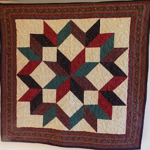 May include: A square quilt featuring a central star pattern composed of dark red, teal, and black fabric pieces. The star is set against a cream-colored background with swirling quilted textures, all enclosed by a dark floral patterned border.