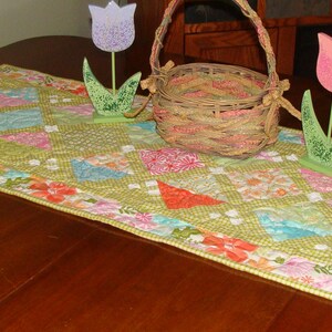 May include: A green and white quilted table runner with a floral border. The runner is decorated with wooden tulips and a wicker basket.