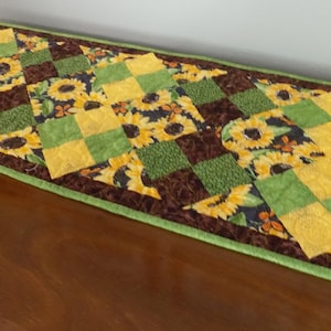 May include: A rectangular table runner with a patchwork design featuring sunflowers, green squares, and brown fabric. The runner has a green border and is placed on a wooden surface.