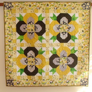 May include: A yellow, black, green, and white quilt with a floral pattern. The quilt is hanging on a wall and has a border of bees and flowers.