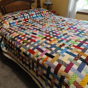 May include: A colorful patchwork quilt with a woven pattern, featuring a variety of fabrics in different colors and textures. The quilt is laid out on a bed with a wooden headboard.
