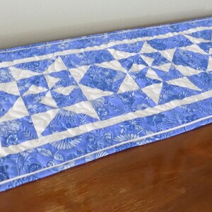 May include: A rectangular quilted table runner featuring a blue and white floral pattern. The design includes a central diamond motif flanked by two star-shaped blocks, all bordered by a solid white frame and a blue floral outer edge.