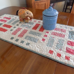 May include: A quilted table runner with a repeating pattern of red and blue rectangles on a cream background. A blue ceramic jar and a decorative elephant figurine sit on the runner. The runner is approximately 12 inches wide.