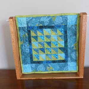 May include: A small, square quilt with a geometric pattern in shades of blue and green, displayed in a wooden frame. The quilt features a patchwork design with triangles and floral accents. The frame is made of light-colored wood.