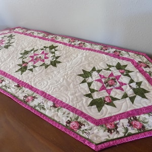 May include: A quilted table runner with a pink and green floral pattern. The runner features a starburst design in the center and is edged with a pink and green floral border.