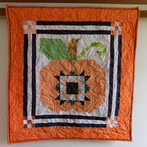 May include: A square quilted wall hanging featuring a central orange pumpkin motif with a green leaf and stem. The pumpkin is decorated with a geometric star pattern in the center. The quilt has a wide orange border with swirling stitching.