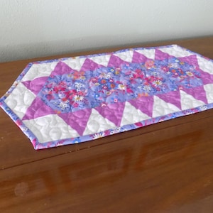 May include: A quilted table runner featuring a central panel of purple fabric with a multi-colored floral print. The runner is bordered by a geometric pattern of white and purple triangles, with overall stippled quilting and a purple floral binding.