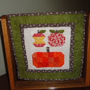 May include: A framed quilted wall hanging with a green and brown border. The center of the hanging features three appliquéd apples, one red, one yellow, and one orange.