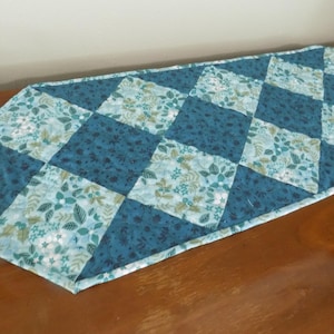 May include: A quilted table runner featuring a diamond-patterned design with alternating fabric squares. The runner displays a color palette of dark teal and light blue, with floral and leaf motifs printed on the fabric surfaces throughout.