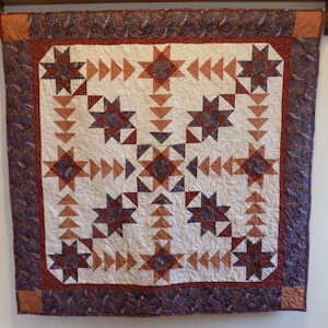 May include: A handmade quilt with a star pattern in shades of red, brown, and cream. The quilt features a central design of stars and triangles, framed by a paisley border. It is displayed on a wooden rod.