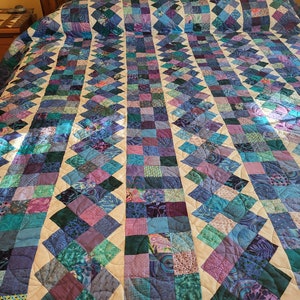 May include: A quilt with a patchwork design featuring squares of various shades of blue, purple, and green. The quilt is sewn together with white fabric and has a curved quilting pattern.