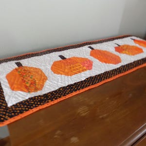 May include: A rectangular quilted table runner featuring a white center panel with five orange pumpkin appliques. The runner is bordered by a black fabric with small orange dots and finished with a bright orange binding along the outer edges.