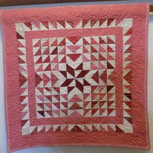 May include: A pink and white quilt with a geometric pattern of triangles and stars. The quilt is quilted with a small, repeating pattern.