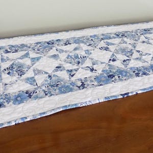 May include: A rectangular table runner with a blue and white floral pattern. The runner features a patchwork design with triangular shapes and a white border. The color palette includes shades of blue and white, creating a soft, inviting aesthetic.