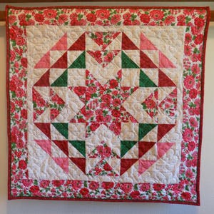 May include: A square quilted wall hanging featuring a central star pattern composed of red floral, solid red, solid green, and solid pink fabric triangles. The quilt has a white background with small dots and a wide border of red floral fabric.