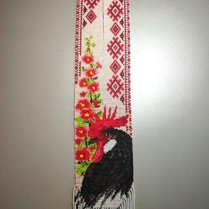 May include: A long, rectangular beaded necklace featuring a black rooster with a bright red comb and wattle, positioned next to a vertical arrangement of pink flowers with green leaves. The upper section displays red and black geometric patterns.