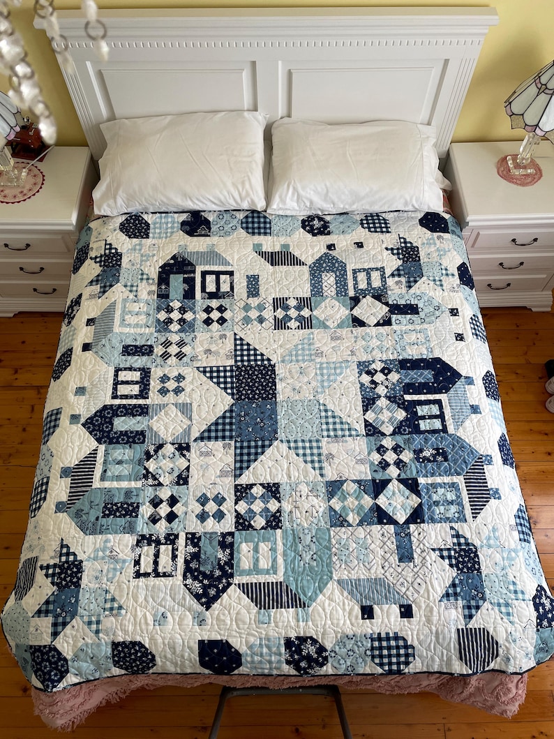 May include: A white and blue patchwork quilt with a variety of patterns, including houses, stars, and geometric shapes. The quilt is laid out on a bed with white pillows.