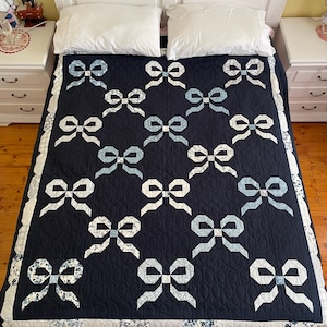 May include: A navy blue quilt with a repeating pattern of white and light blue bows. The quilt has a scalloped edge with a floral print border. The bed is made with white pillows and a white headboard.