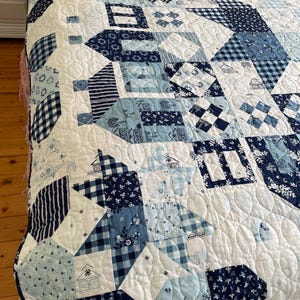 May include: A white quilt with a blue and white patchwork design. The quilt is made up of various geometric shapes and patterns, including squares, triangles, and circles. The quilt is quilted with a wavy pattern.