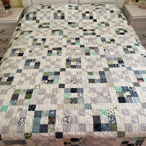 Può includere: Una trapunta patchwork nei toni del grigio, bianco, blu e verde copre un letto. La trapunta presenta un motivo ripetuto di quadrati e rettangoli, con vari disegni di tessuto floreale e geometrico. La trapunta è un elemento di biancheria da letto accogliente e decorativo.