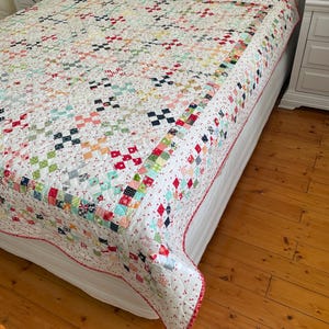 May include: A white quilt with a patchwork design featuring various colors and patterns, including red, pink, blue, and green. The quilt is on a white bed with a white headboard and is surrounded by a wooden floor.