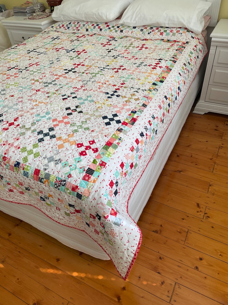 May include: A white quilt with a patchwork design featuring various colors and patterns, including red, pink, blue, and green. The quilt is on a white bed with a white headboard and is surrounded by a wooden floor.