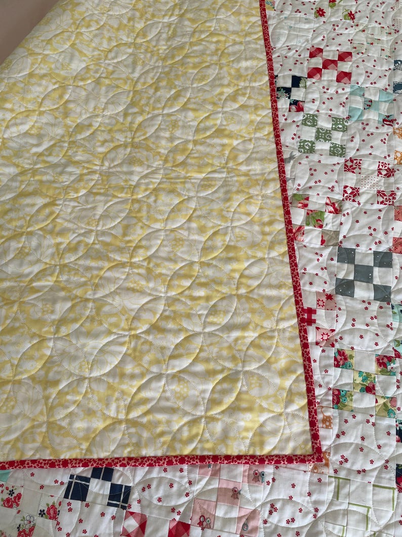 May include: A yellow and white quilt with a red binding. The quilt is quilted with a pattern of curved lines. The back of the quilt is made of patchwork squares in various colors and patterns.