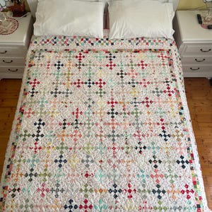 May include: A white quilt with a patchwork design featuring a variety of colorful fabrics. The quilt is made up of small squares and rectangles, and it has a quilted pattern of small circles. The quilt is on a bed with white pillows and a white headboard.