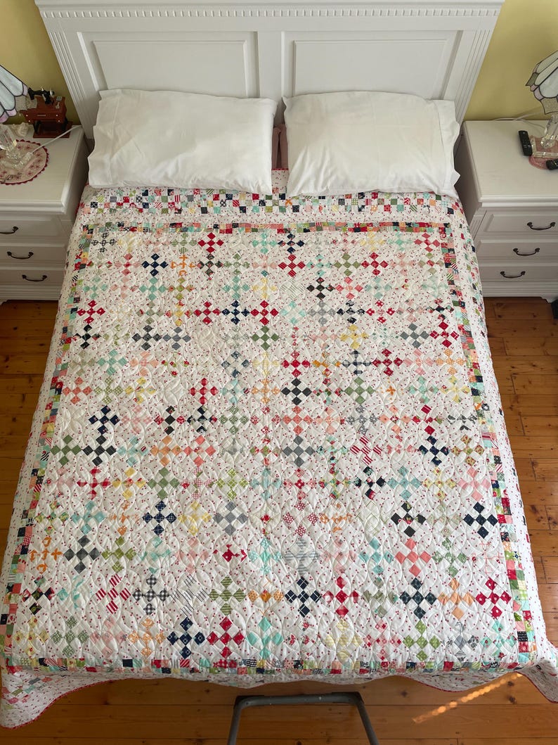 May include: A white quilt with a patchwork design featuring a variety of colorful fabrics. The quilt is made up of small squares and rectangles, and it has a quilted pattern of small circles. The quilt is on a bed with white pillows and a white headboard.