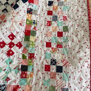 May include: A white quilt with a red and white checkered pattern on the left side and a patchwork pattern on the right side. The quilt is quilted with a pattern of small red flowers and curved lines.