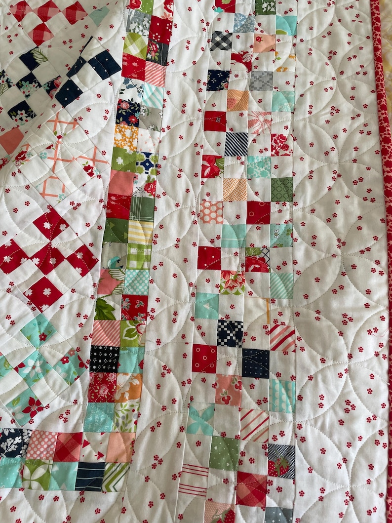 May include: A white quilt with a red and white checkered pattern on the left side and a patchwork pattern on the right side. The quilt is quilted with a pattern of small red flowers and curved lines.