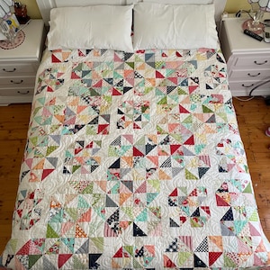 May include: A handmade quilt with a patchwork design of colorful triangles in various patterns and colors, including red, blue, green, and floral prints. The quilt is on a white bed with white pillows and a white headboard. The quilt is a cozy and decorative bedding item.