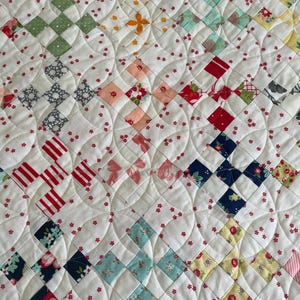 May include: A white quilt with a patchwork design featuring various floral and geometric patterns in shades of red, blue, green, and yellow. The quilt is quilted with a curved design.