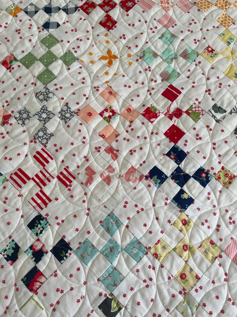 May include: A white quilt with a patchwork design featuring various floral and geometric patterns in shades of red, blue, green, and yellow. The quilt is quilted with a curved design.