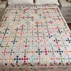 Puede incluir: Una colcha blanca con un diseño de patchwork que presenta una variedad de colores y patrones, incluyendo rojo, azul, verde y amarillo. La colcha está hecha de cuadrados pequeños y está acolchada con hilo blanco. La colcha está sobre una cama con sábanas y almohadas blancas.