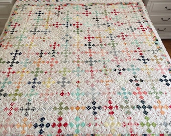 May include: A white quilt with a patchwork design featuring a variety of colors and patterns, including red, blue, green, and yellow. The quilt is made up of small squares and is quilted with a white thread. The quilt is on a bed with white sheets and pillows.