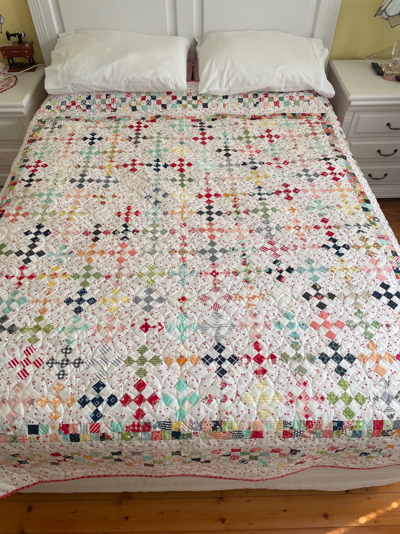 May include: A white quilt with a patchwork design featuring a variety of colors and patterns, including red, blue, green, and yellow. The quilt is made up of small squares and is quilted with a white thread. The quilt is on a bed with white sheets and pillows.